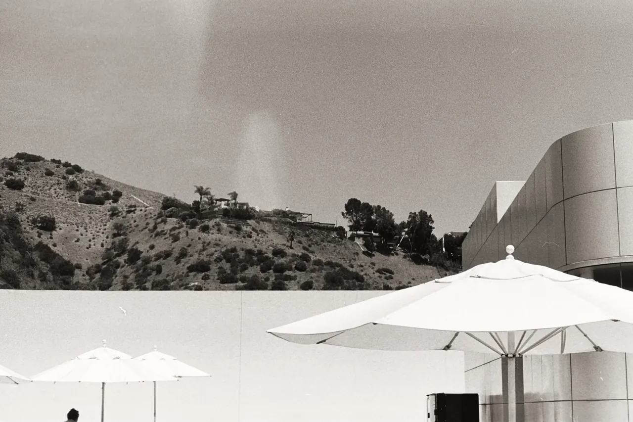 Black-and-white photo of a hillside with palm trees and a modernist curved building, striped beach umbrellas in the foreground