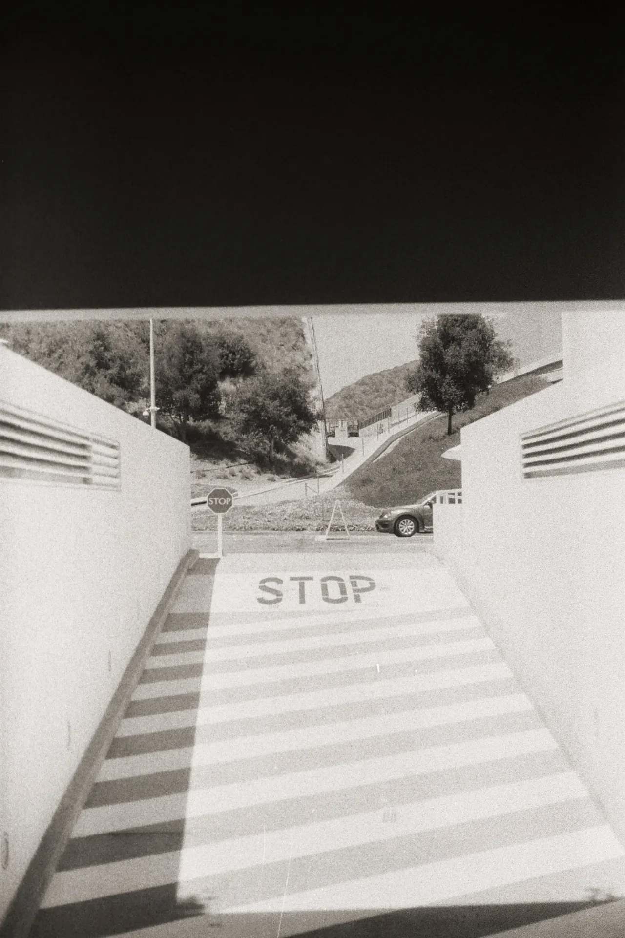 Black-and-white photo looking down a covered ramp toward a STOP sign and a hillside road