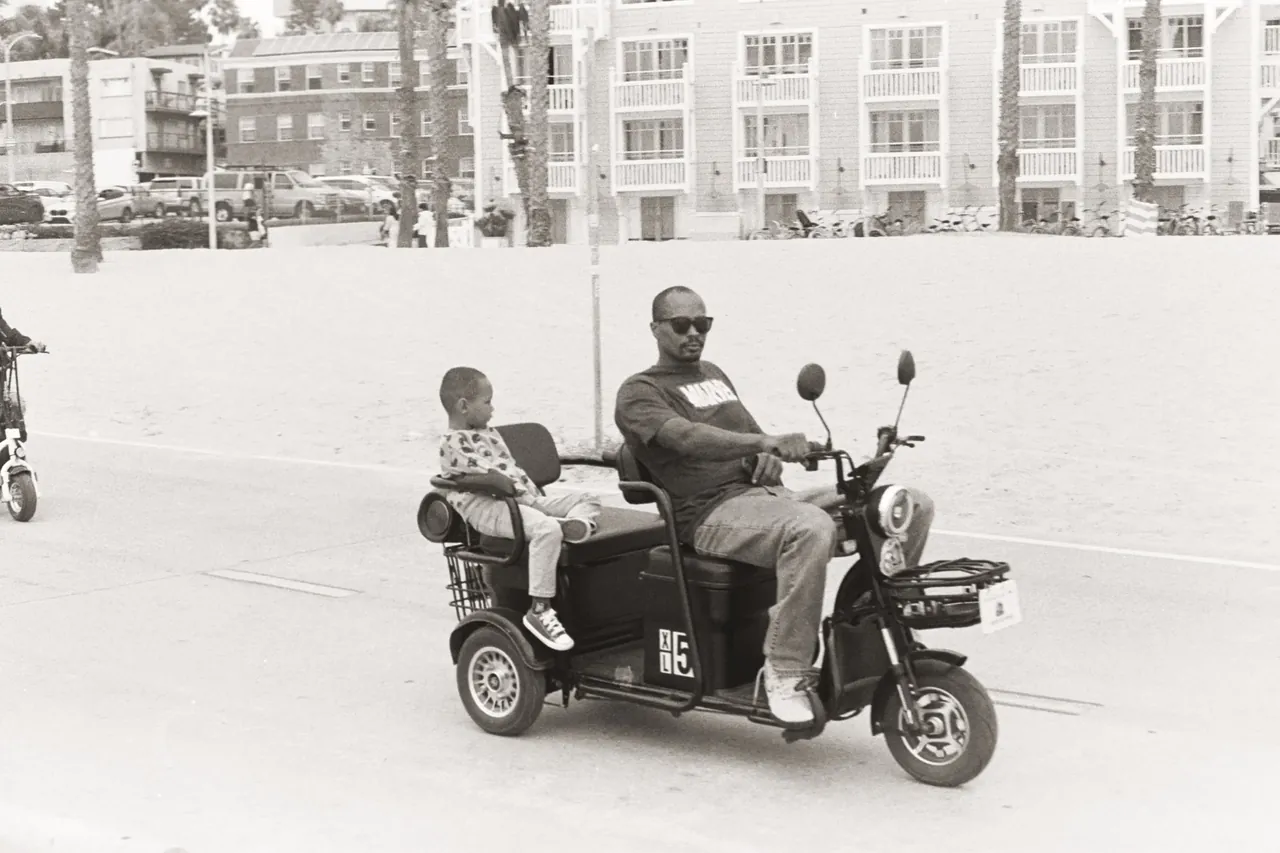 Black-and-white photo of a man and child riding a three-wheeled mobility scooter along a beachfront boardwalk