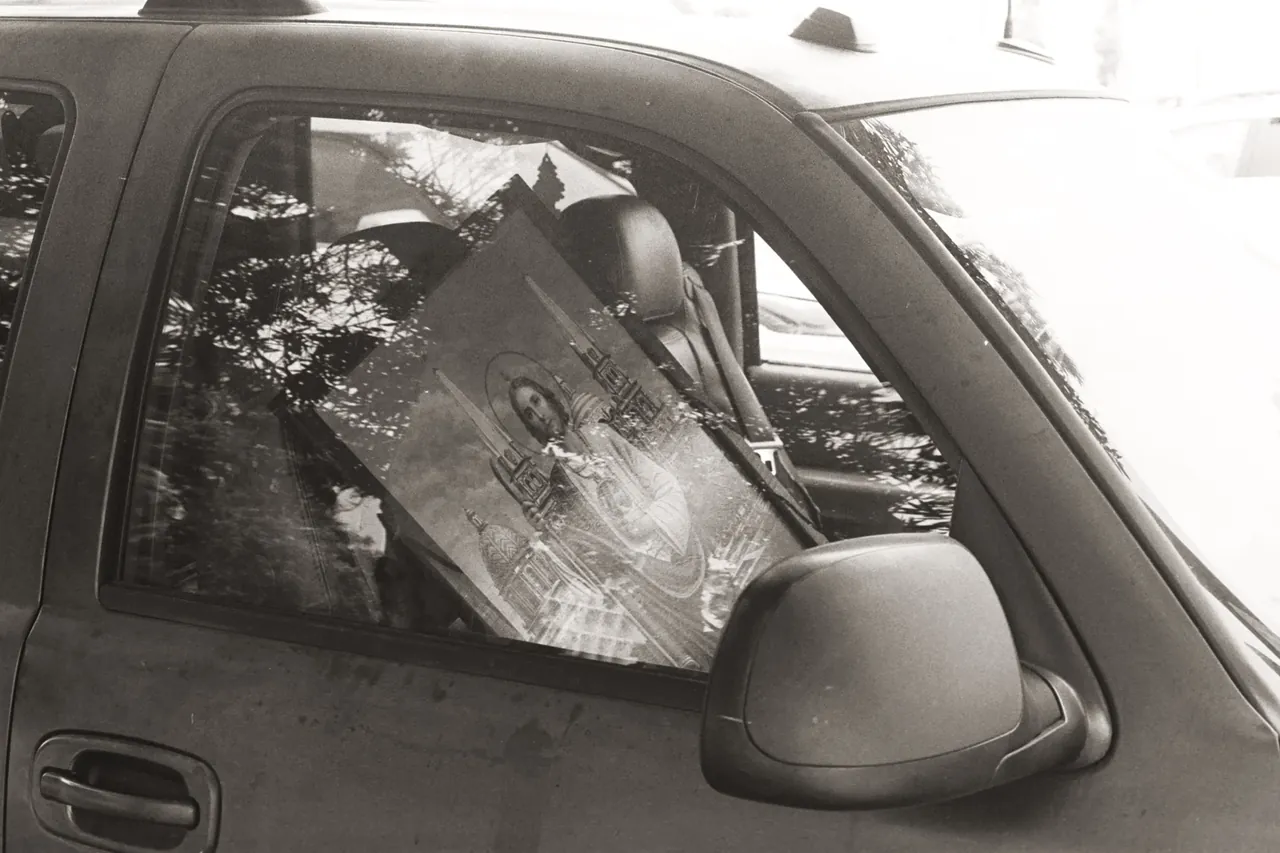Black-and-white photo of a religious icon painting propped in a car’s back window, tree branches reflected in the glass