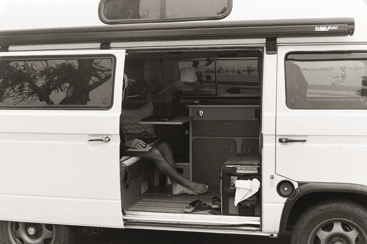 Black-and-white photo of a man sitting barefoot with a laptop in the open side door of a VW camper van