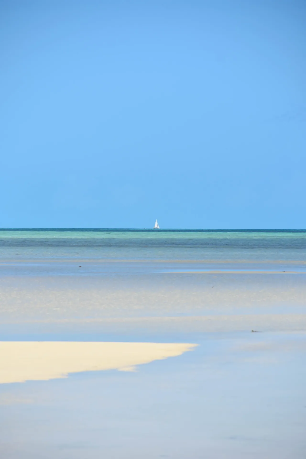 A minimalist seascape with a single white sailboat on the horizon above a pale tidal flat